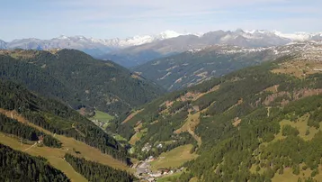 Panoramablick auf ein weites Alpental mit bewaldeten Hängen und schneebedeckten Berggipfeln in der Ferne.