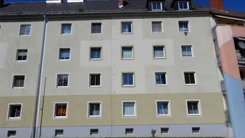 Mehrfamilienhaus mit heller Fassade, vielen Fenstern und einem Satteldach unter blauem Himmel.
