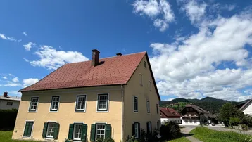 Traditionelles gelbes Haus mit rotem Ziegeldach und grünen Fensterläden unter blauem Himmel.