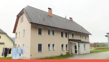 Mehrfamilienhaus mit heller Fassade und Satteldach in ländlicher Umgebung, umgeben von Grünflächen.