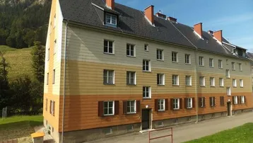 Mehrfamilienhaus mit gestreifter Fassade und Satteldach vor einer malerischen Bergkulisse.