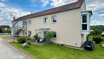 Mehrfamilienhaus mit heller Fassade, kleinem Vorgarten und verglastem Anbau unter blauem Himmel.