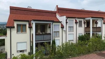 Mehrfamilienhaus mit rotem Ziegeldach und Balkonen in ländlicher Umgebung.