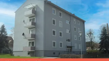 Mehrfamilienhaus mit grauer Fassade, mehreren Balkonen und umgebender Grünfläche unter blauem Himmel.