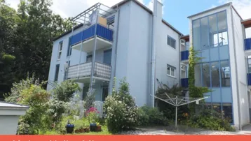 Mehrfamilienhaus mit mehreren Balkonen und umgebender Grünfläche unter blauem Himmel.