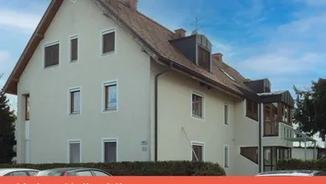 Gebäudeansicht eines Wohnhauses mit heller Putzfassade, Ziegeldach, Gauben und Rollläden an den Fenstern.