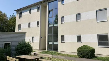 Außenansicht eines modernen Mehrfamilienhauses mit zentraler Glasfassade und Sitzgelegenheiten im Grünen.