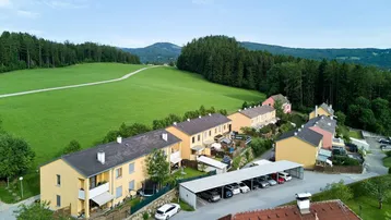 Luftaufnahme einer Wohnsiedlung mit Reihenhäusern, umgeben von grünen Feldern und Wäldern.