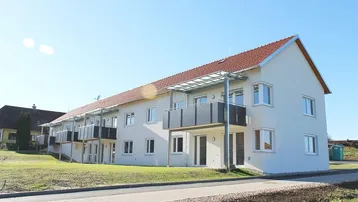 Modernes Mehrfamilienhaus mit weißer Fassade, roten Ziegeldächern und Balkonen in ländlicher Umgebung.