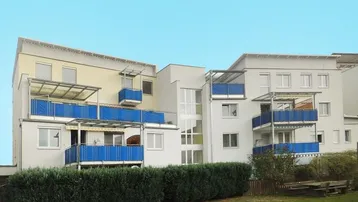 Modernes Mehrfamilienhaus mit blauen Balkonen und gepflegter Grünanlage im Vordergrund.