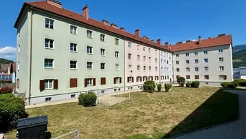 Mehrfamilienhaus mit hellgrüner Fassade, roten Ziegeldächern und einem gemeinschaftlichen Rasenhof unter blauem Himmel.
