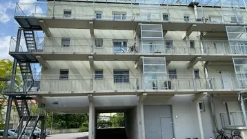 Modernes Mehrfamilienhaus mit mehreren Balkonen, Außentreppe und Tiefgaragenzufahrt, umgeben von Grünflächen.