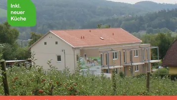 Mehrfamilienhaus in ländlicher Umgebung mit Apfelbäumen im Vordergrund und grünen Hügeln im Hintergrund.