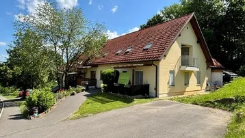 Gut gepflegtes Einfamilienhaus mit gelber Fassade, rotem Satteldach und kleinem Balkon, umgeben von viel Grün.