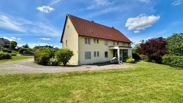 Freistehendes Einfamilienhaus mit Satteldach, großem Rasen und gepflasterter Auffahrt an einem sonnigen Tag.