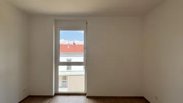 Helles, leeres Zimmer mit Laminatboden und einem großen Fenster mit Blick nach draußen.