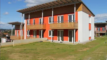 Modernes Mehrfamilienhaus mit leuchtend oranger Fassade, Balkonen und großem Gartenbereich unter blauem Himmel.