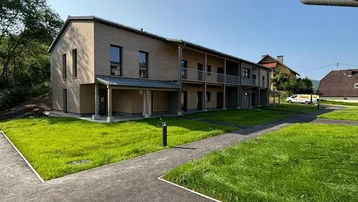 Modernes Mehrfamilienhaus mit Holzfassade, Balkonen und gepflegten Grünflächen unter blauem Himmel.