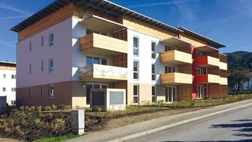 Modernes Mehrfamilienhaus mit Holzverkleidung, großen Fenstern und mehreren Balkonen in sonniger Hanglage.