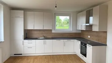 Neue, helle Einbauküche mit weißen Schränken, dunkler Arbeitsplatte und Holzoptik-Rückwand.