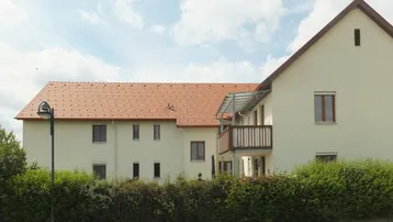Ansicht eines Mehrfamilienhauses mit heller Fassade, Satteldach und einem Balkon.