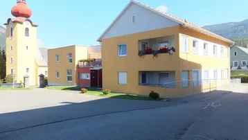 Gebäudeansicht mit gelber Fassade und angrenzendem Kirchturm unter blauem Himmel.