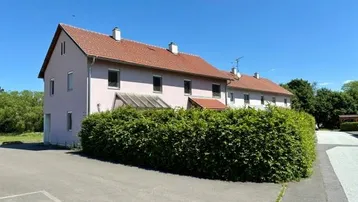 Rosa gestrichenes Wohngebäude mit rotem Ziegeldach und gepflegter Hecke vor dem Eingangsbereich.