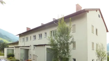 Mehrfamilienhaus mit heller Fassade und Satteldach, umgeben von grüner Landschaft und Bergen.