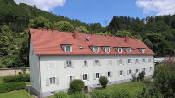 Mehrfamilienhaus mit rotem Mansarddach und hellgrüner Fassade, umgeben von grünen Hügeln und Bäumen.