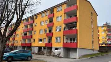 Mehrfamilienhaus mit gelber Fassade und roten Balkonen in einer Wohngegend.