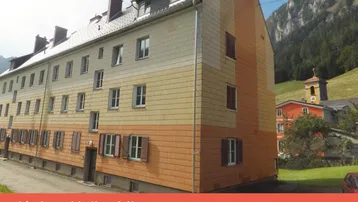 Älteres Mehrfamilienhaus mit farbiger Fassade und Blick auf die umliegenden Berge und grüne Landschaft.