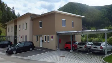 Modernes Gebäude mit Parkplätzen und Carport vor einer bewaldeten Bergkulisse.