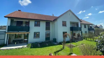 Mehrfamilienhaus mit gepflegtem Garten, Balkonen und einer Terrasse unter blauem Himmel.