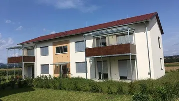 Modernes Mehrfamilienhaus mit Balkonen und rotem Dach in ländlicher Umgebung.