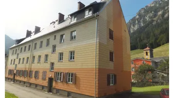 Mehrfamilienhaus in den Bergen mit farbiger Fassade, Satteldach und Fensterläden, umgeben von grüner Natur.