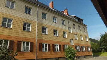 Mehrfamilienhaus mit gestreifter Fassade, traditionellen Fensterläden und grüner Umgebung unter blauem Himmel.