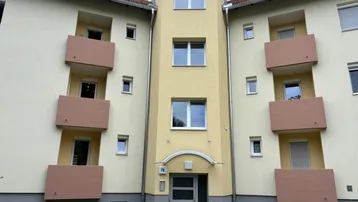Mehrfamilienhaus mit gelber Fassade, braunen Balkonen und rotem Ziegeldach in einer Wohngegend.