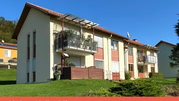 Mehrfamilienhaus mit mehreren Balkonen und gepflegter Grünfläche in ländlicher Umgebung, unter blauem Himmel.