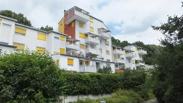 Mehrstöckiges Wohngebäude mit zahlreichen Balkonen und gelben Fensterläden, umgeben von viel Grün.