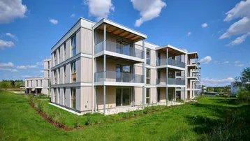 Modernes Mehrfamilienhaus mit hellen Fassaden und Balkonen, umgeben von grüner Landschaft unter blauem Himmel.