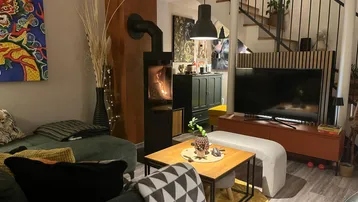 Gemütliches Wohnzimmer mit modernem Holzofen, Sofa und Fernseher, Treppe im Hintergrund.