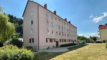 Mehrstöckiges Wohngebäude mit heller Fassade, braunen Fensterläden und gepflegter Grünfläche unter blauem Himmel.