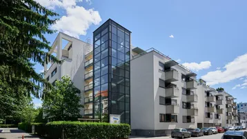 Modernes Mehrfamilienhaus mit Glasaufzug und Balkonen an einer begrünten Straße.