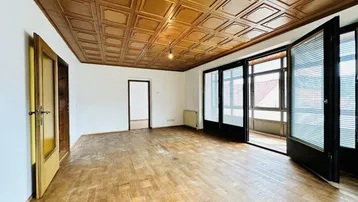 Geräumiges Wohnzimmer mit Holzkassettendecke, Parkettboden und Zugang zu einem verglasten Balkon.