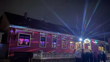 Fassade eines Gebäudes bei Nacht, beleuchtet mit bunten Lichtstrahlen und einer Gruppe von Menschen davor.