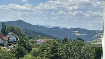 Malerischer Ausblick auf grüne Hügel, Bäume und Häuser unter einem bewölkten Himmel.