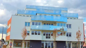 Modernes Mehrfamilienhaus mit weißer Fassade, orangefarbenen Akzenten und blauen Glasbalkonen unter bewölktem Himmel.