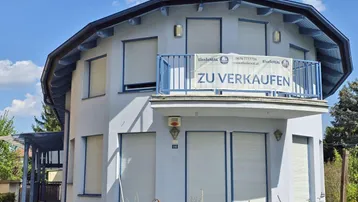 Rundes Haus mit hellblauer Fassade und weißen Rollläden, ein "Zu Verkaufen" Banner hängt am Balkon.
