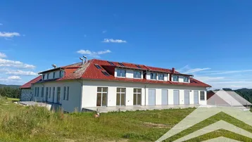 Großes Gebäude mit rotem Ziegeldach und weißer Fassade in ländlicher Umgebung unter blauem Himmel.