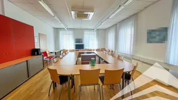 Großer Konferenzraum mit U-förmigem Tisch, vielen Stühlen und hellen Vorhängen an den Fenstern.